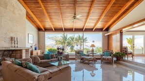 A spacious living area with a high ceiling, wooden beams, and large windows offering a view of palm trees. - Posada El Quijote (San Rafael)
