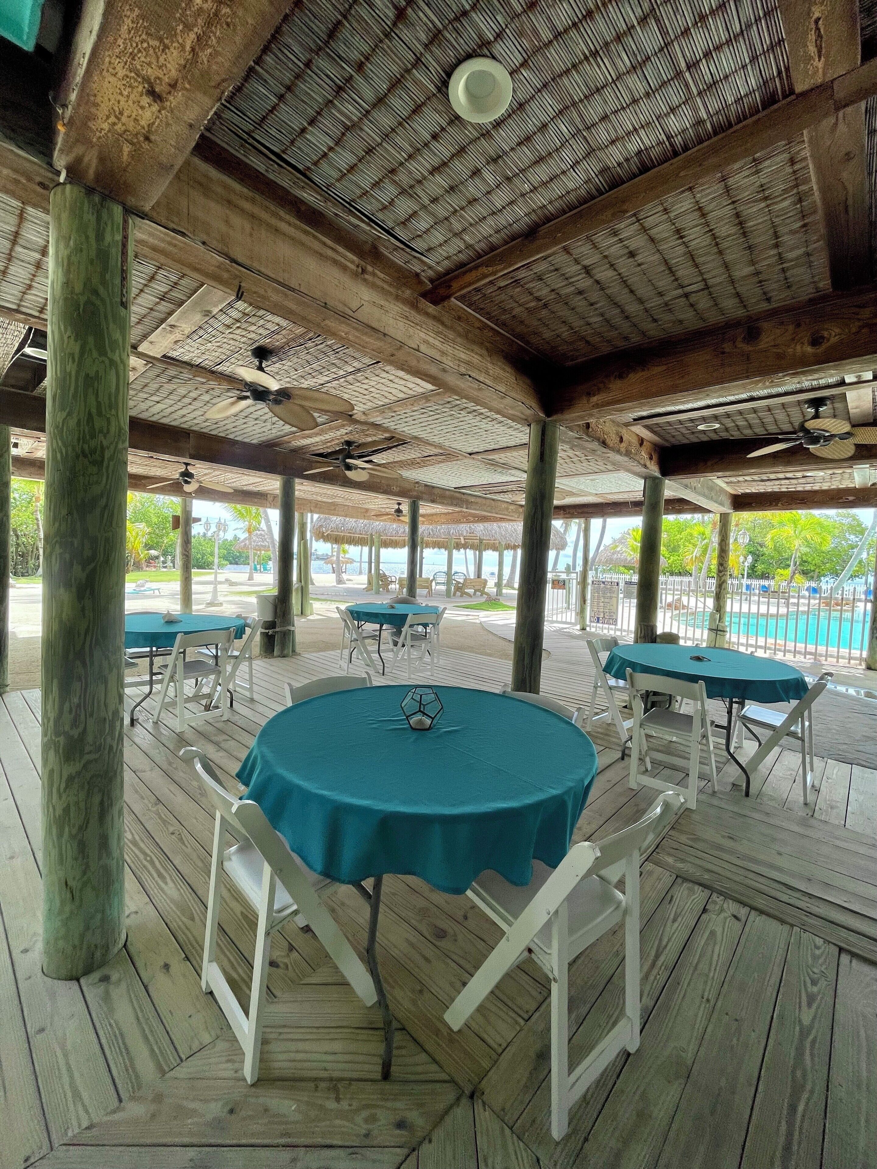 Un espace extérieur abrité, doté de tables et de chaises, avec une piscine en arrière-plan et un plafond en bois agrémenté de ventilateurs.
