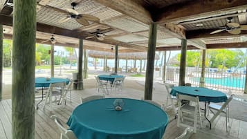 Un espace extérieur abrité, doté de tables et de chaises, avec une piscine en arrière-plan et un plafond en bois agrémenté de ventilateurs.