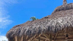 A wooden deck with a thatched roof structure, wooden chairs, and a table, overlooking a tropical landscape with palm trees and a clear sky.