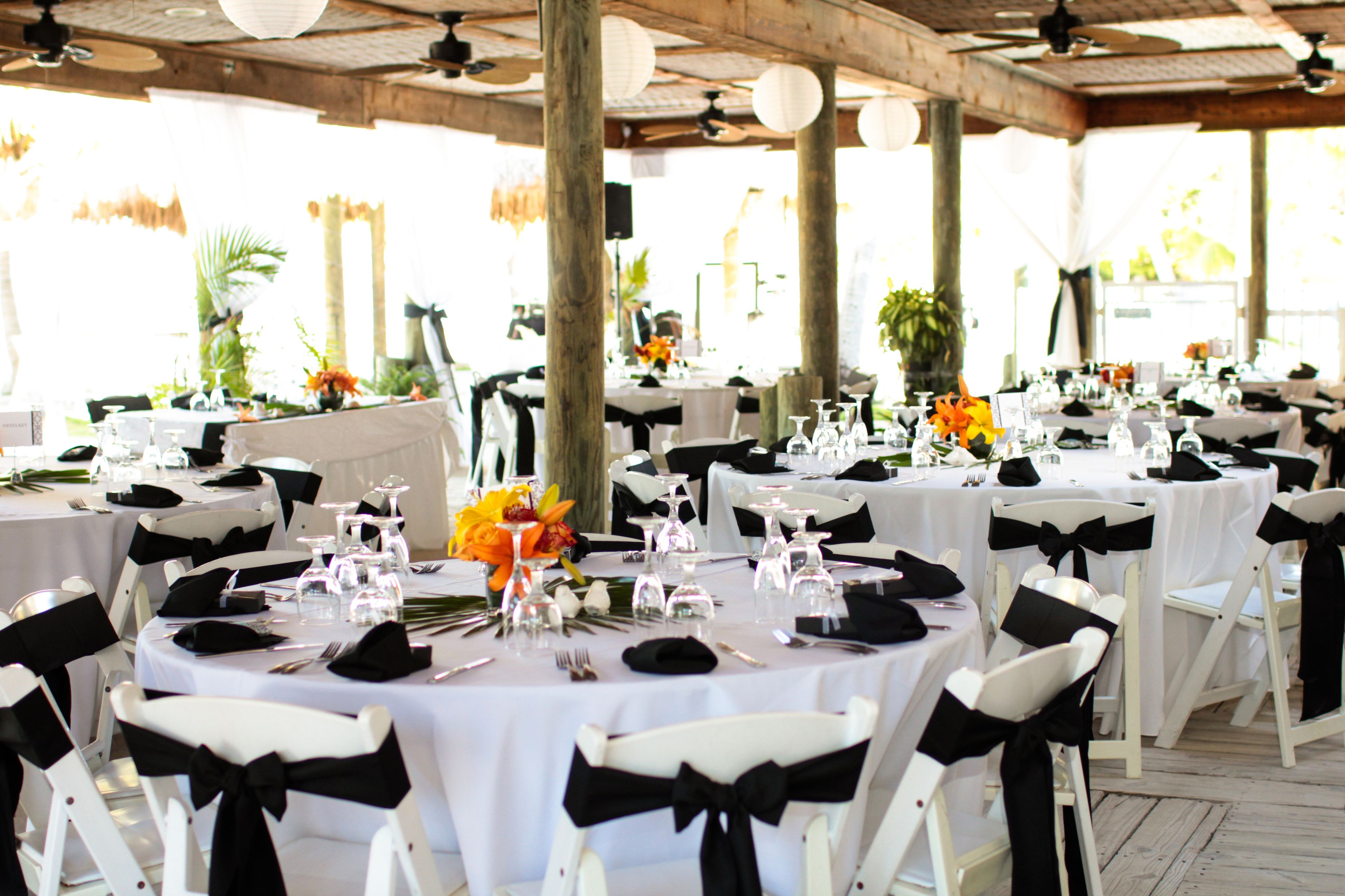 Un salón de banquetes con mesas redondas, sillas blancas y cintas negras. Las mesas están dispuestas con cristalería, platos y cubertería.