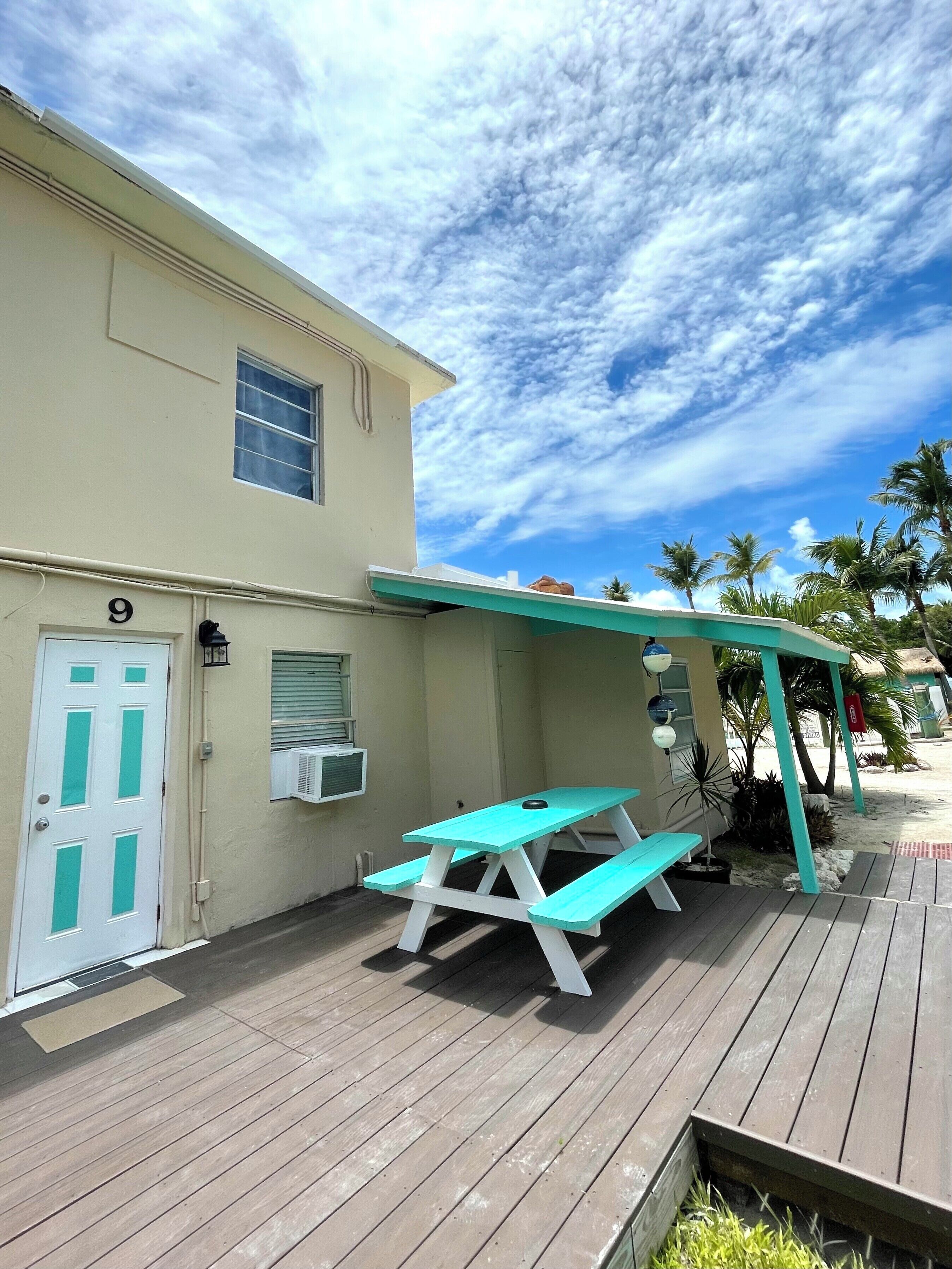 A turquoise picnic table and bench set on a wooden deck, adjacent to a building with a white door and window, under a partly cloudy sky.