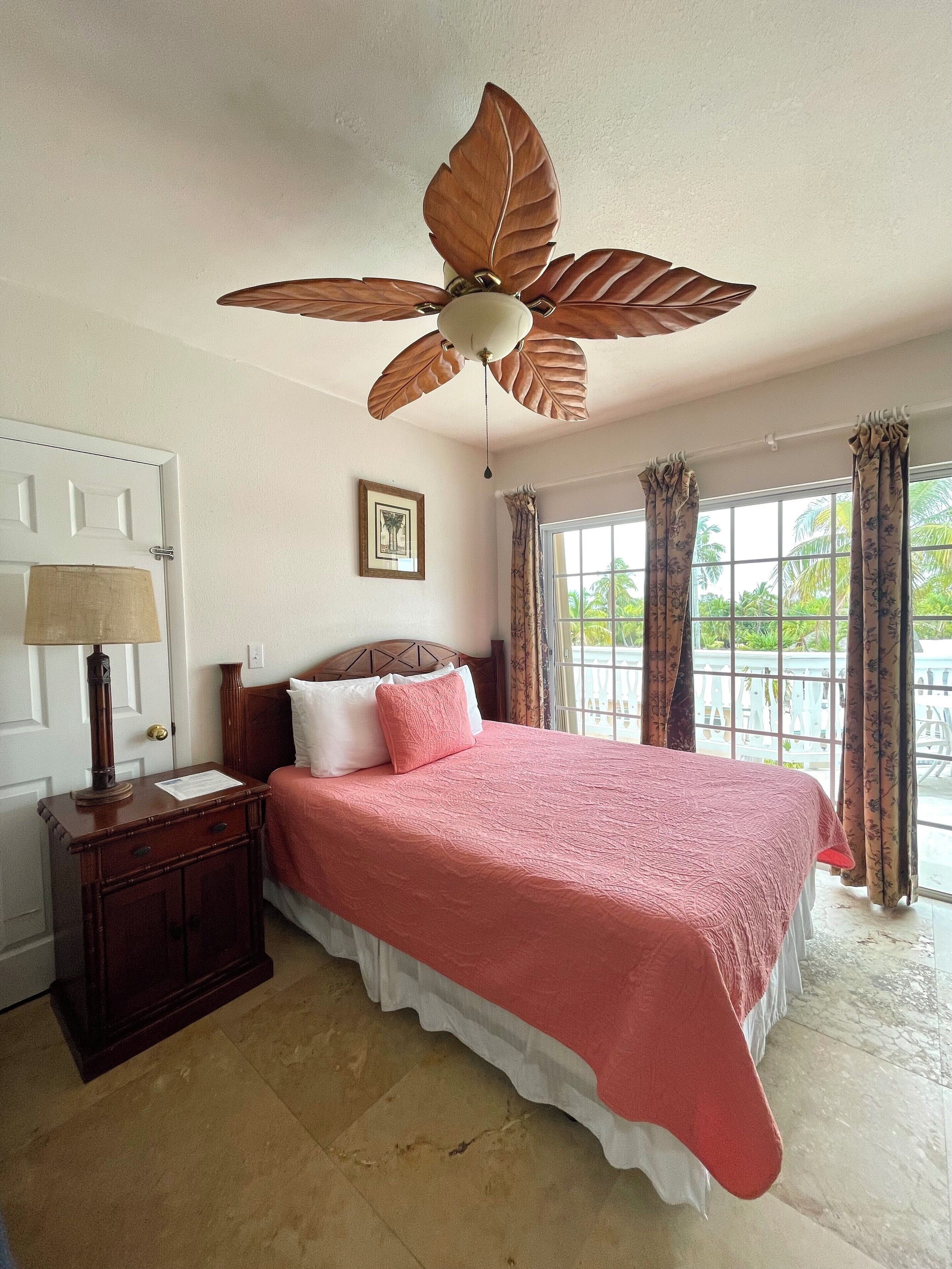 A bedroom with a large bed, a wooden nightstand, a ceiling fan, a window with curtains, and a door.