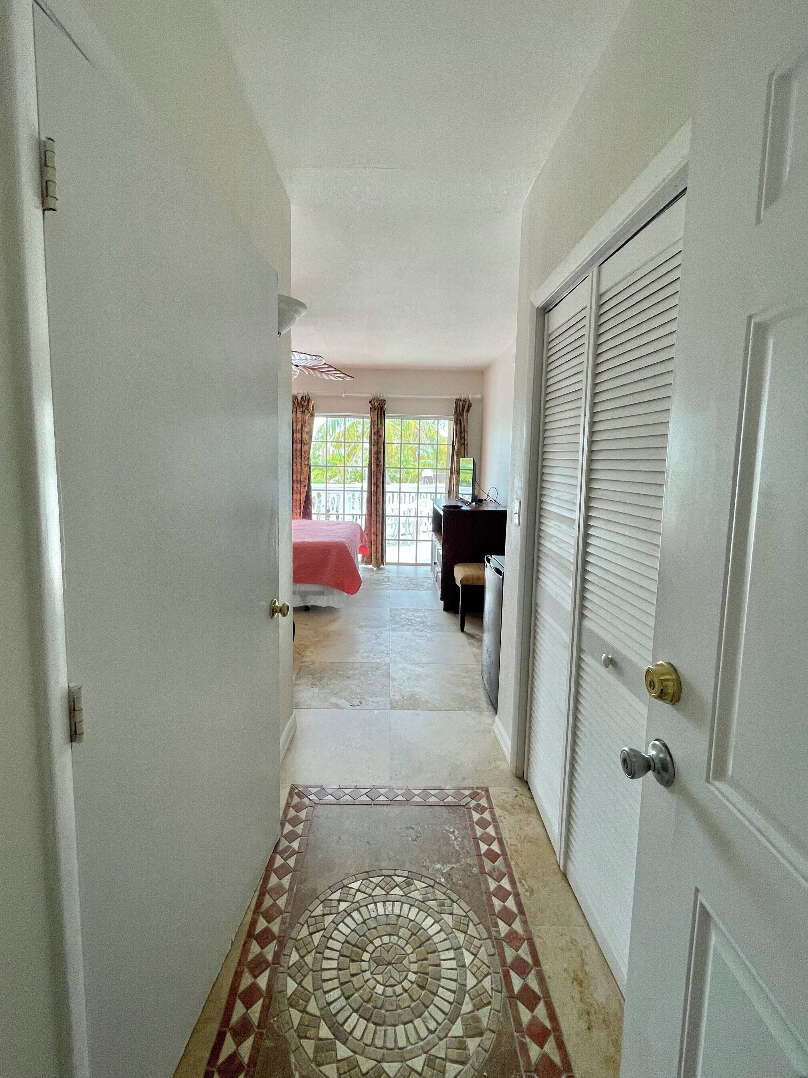 A hallway leading to a bedroom with a bed, a desk, and a window with curtains.