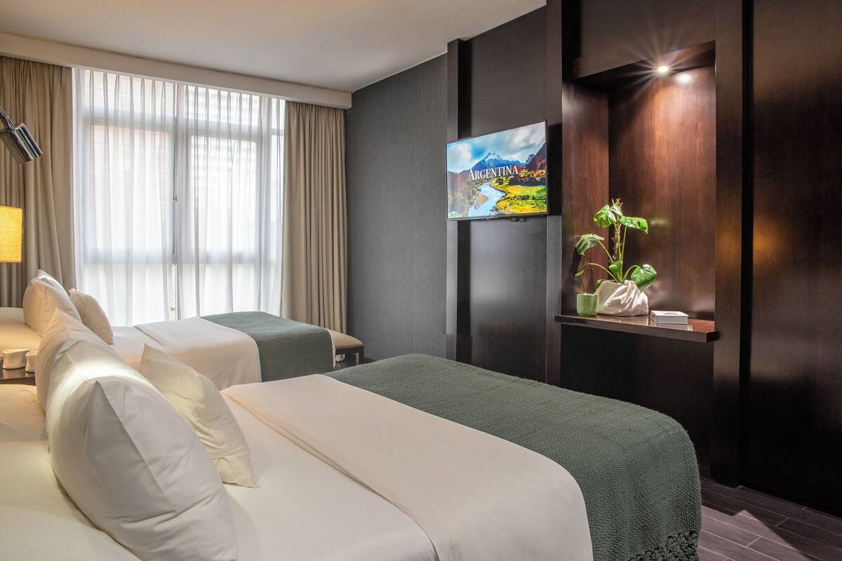 A hotel room with two beds, a large window with curtains, a television on the wall, and a small plant on a shelf.