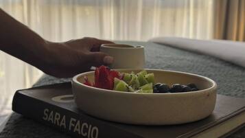 Uma tigela de frutas sobre um livro intitulado "SARA FACIO", perto de uma janela com persianas.