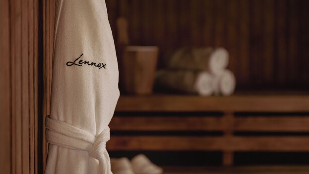 A white bathrobe with the name 'Lennox' embroidered on it, hanging on a wooden wall in a room with a wooden bench and a pair of shoes in the background. - Lennox Hotel Buenos Aires (Buenos Aires)