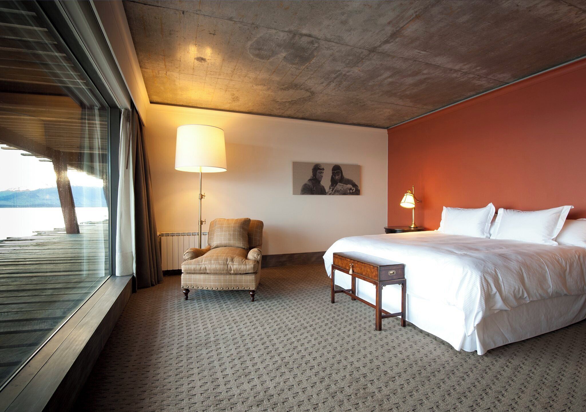 Habitación de hotel con cama, una silla, lámpara, un cuadro en la pared y ventana con vistas a las montañas.
