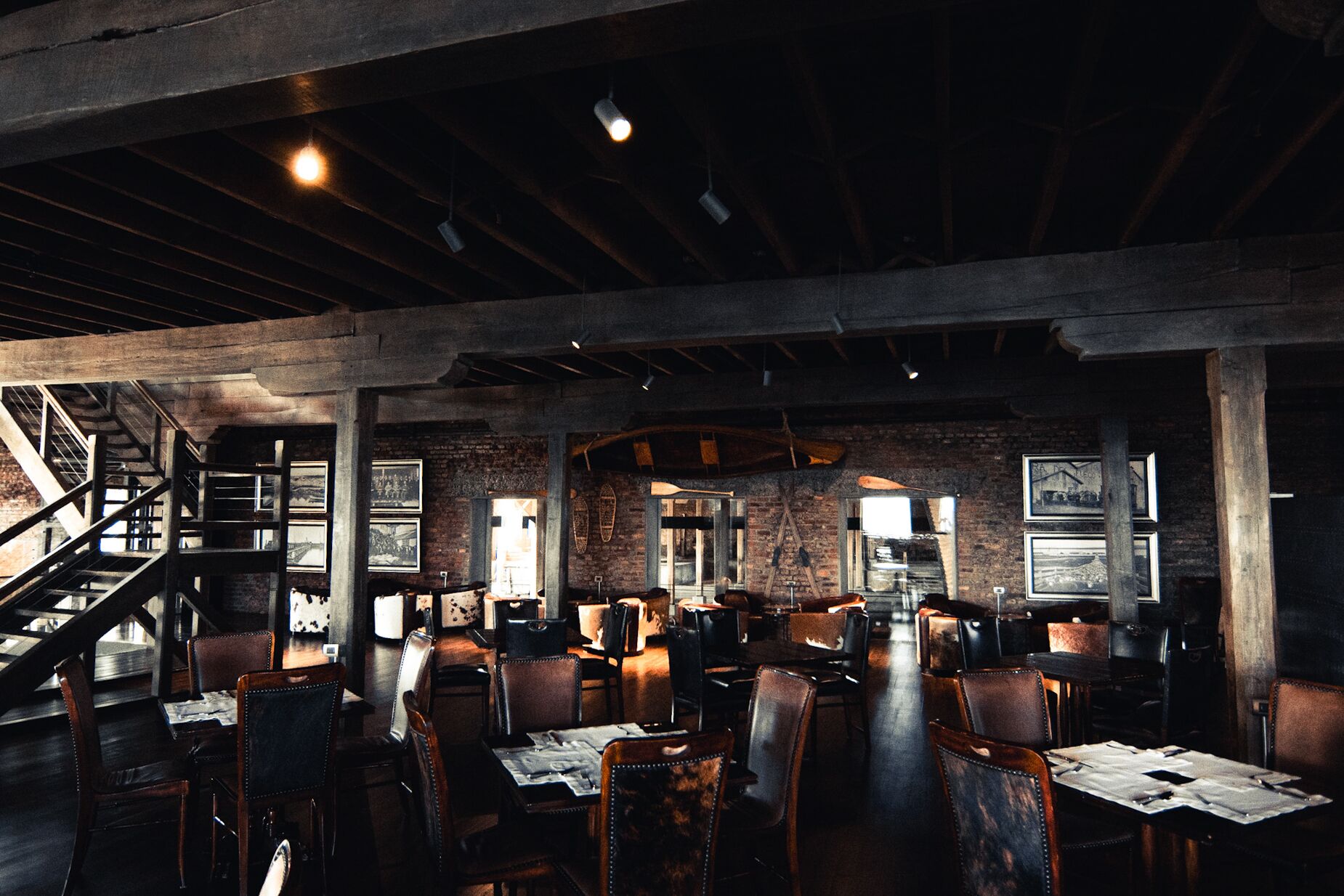 A restaurant with exposed brick walls, wooden beams, and a staircase leading to an upper level. Tables are set with menus and chairs.