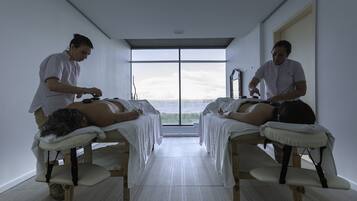 Un cuarto de tratamientos con dos clientes acostados en camillas, un terapeuta atendiendo a cada uno, y un ventanal que ofrece vistas al mar.