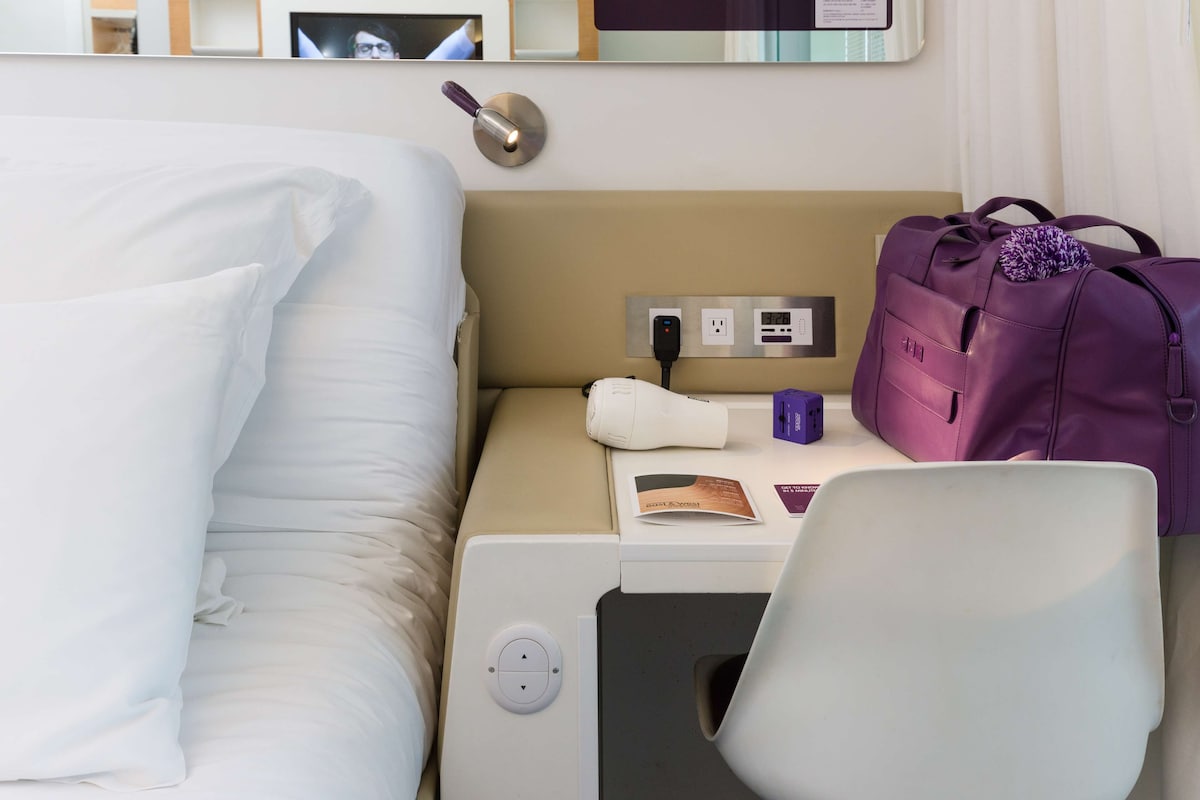 A hotel room with a bed, a bedside table with a lamp, a power outlet, a purple bag, and a chair.