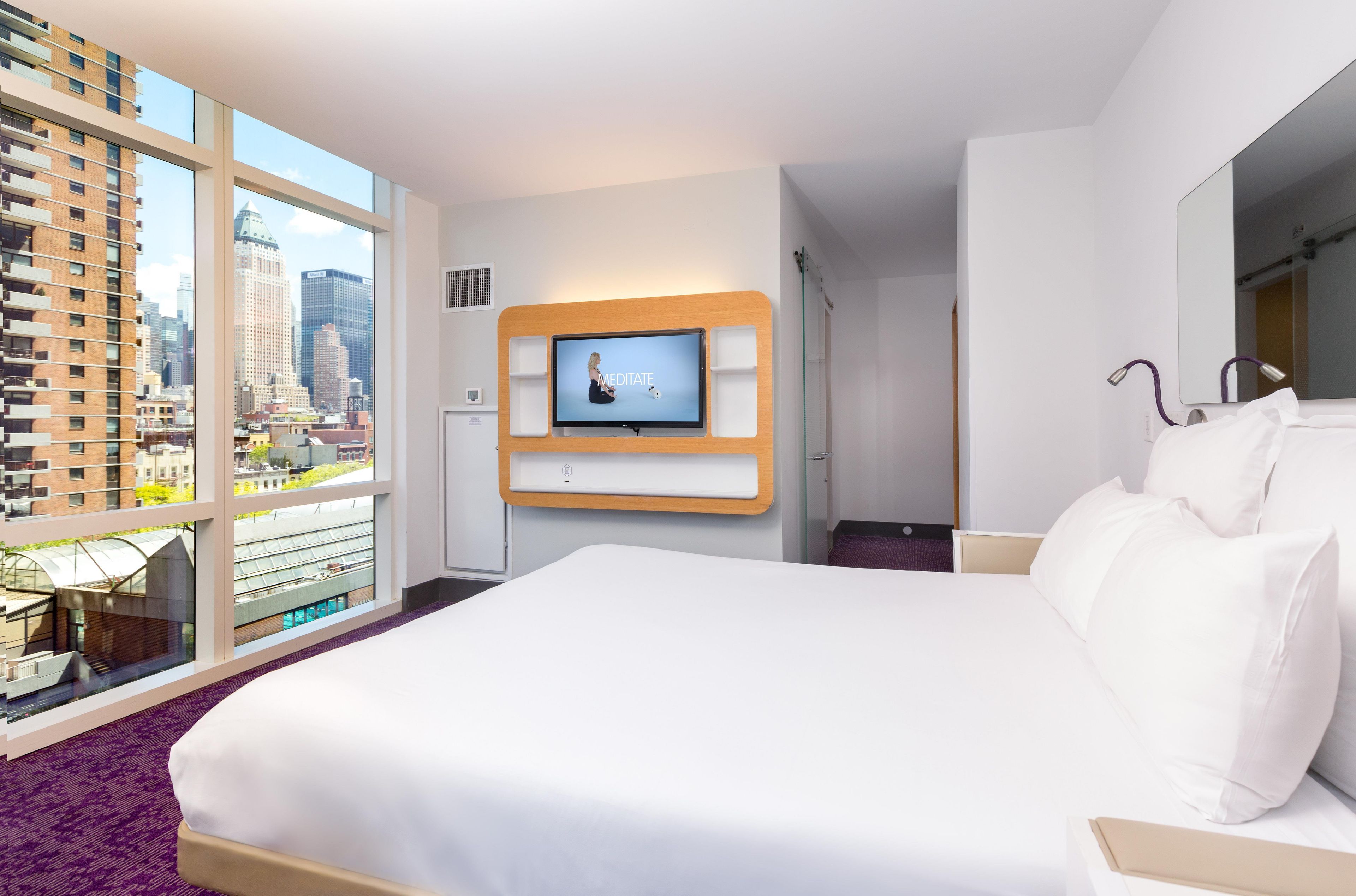 Habitación de hotel con una cama grande, un televisor montado en la pared y vistas de la ciudad a través de amplias ventanas.