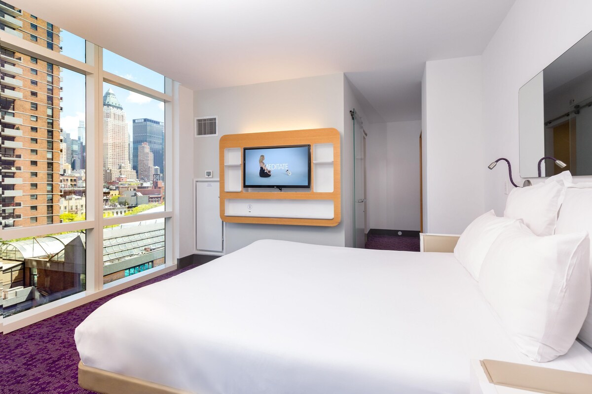 Camera d'albergo con un letto grande, una televisione a parete e vista sulla città attraverso ampie finestre.