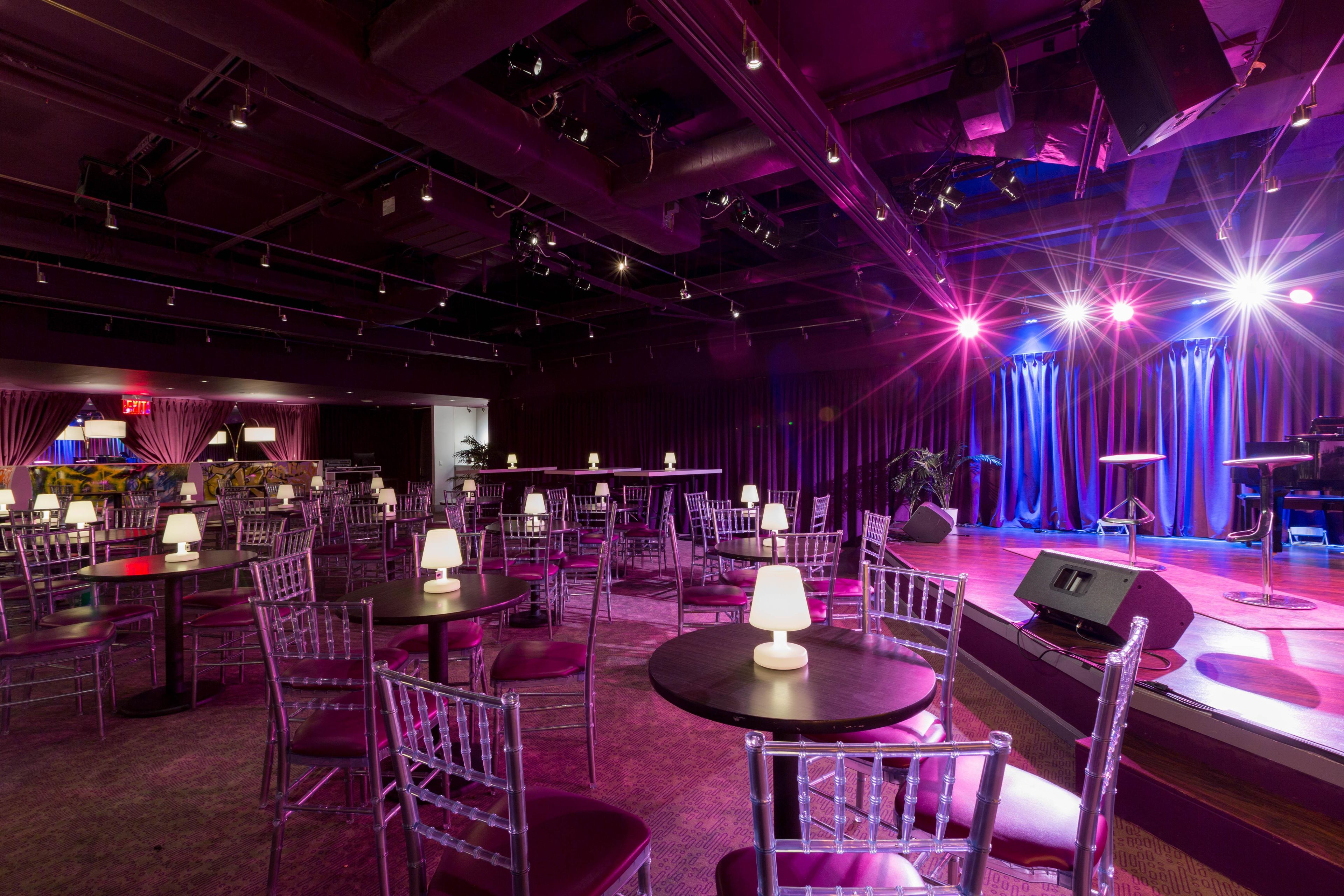 A spacious venue with multiple round tables and chairs, a stage with curtains, and overhead lighting.
