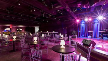 A spacious venue with multiple round tables and chairs, a stage with curtains, and overhead lighting.