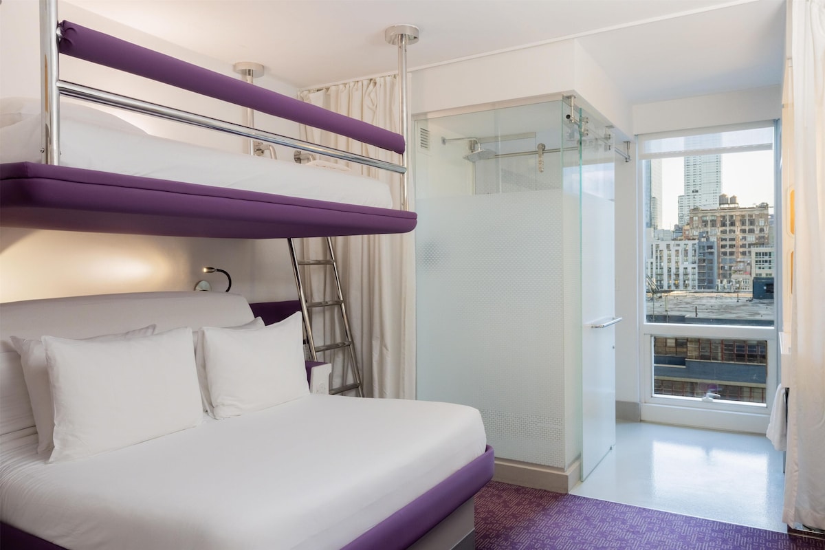 A hotel room with a bed, a ladder, a shower, and a city view.