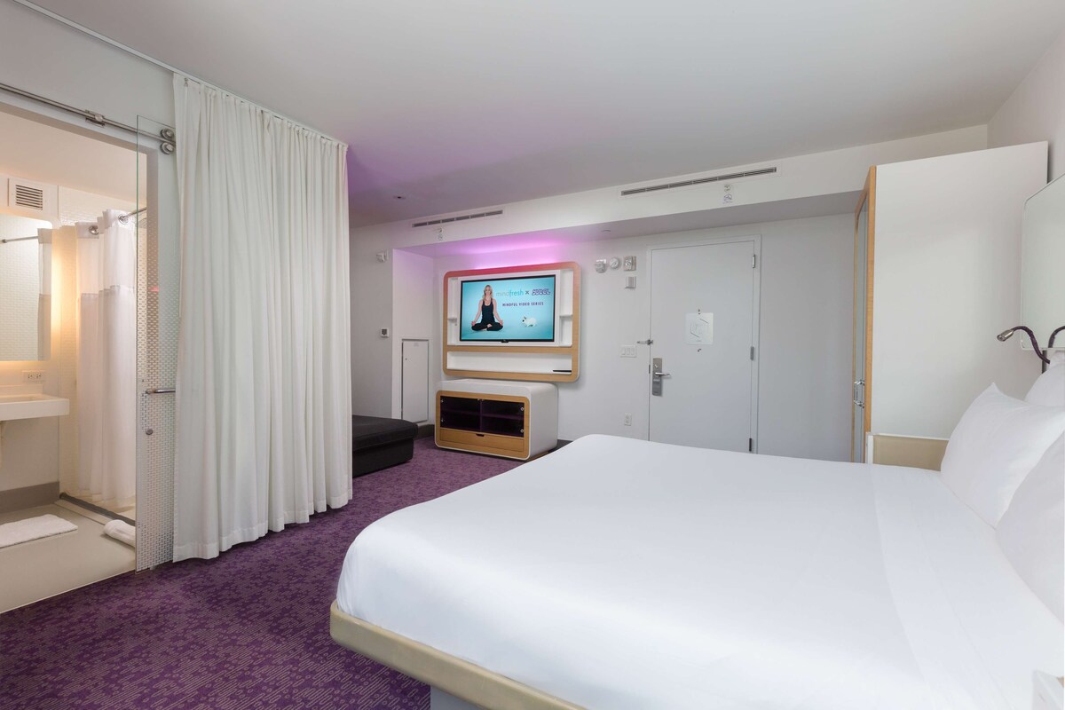 A hotel room with a large bed, a television mounted on the wall, a shower area, and a purple carpet.