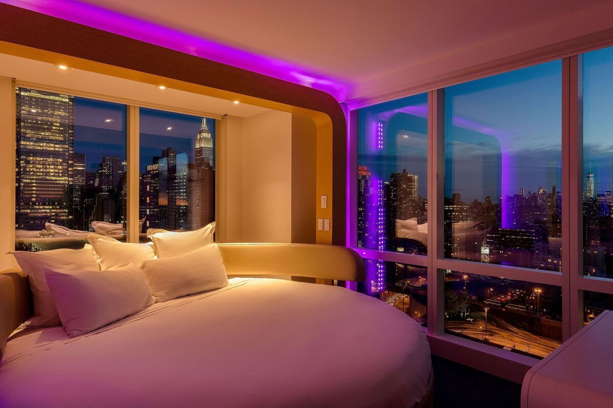 Camera d'albergo con un letto grande, vista sulla città e illuminazione ambientale viola.