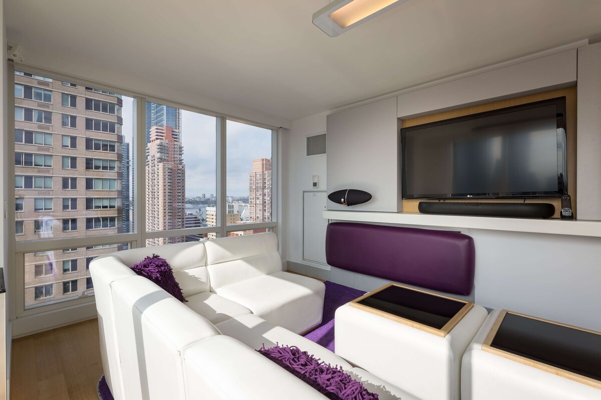 A modern living room with a large window, white leather sofa, purple cushions, and a mounted TV.