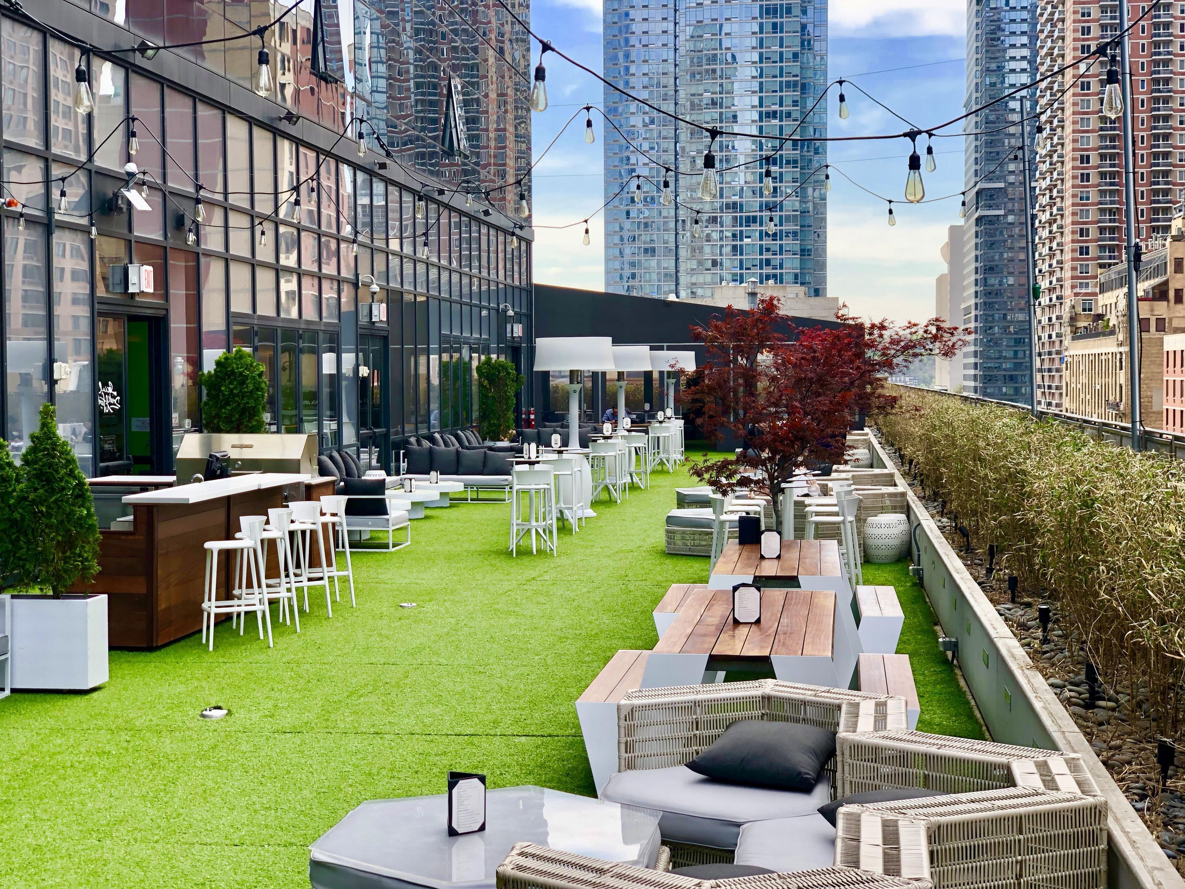 An outdoor seating area with tables and chairs, surrounded by greenery and a view of tall buildings.