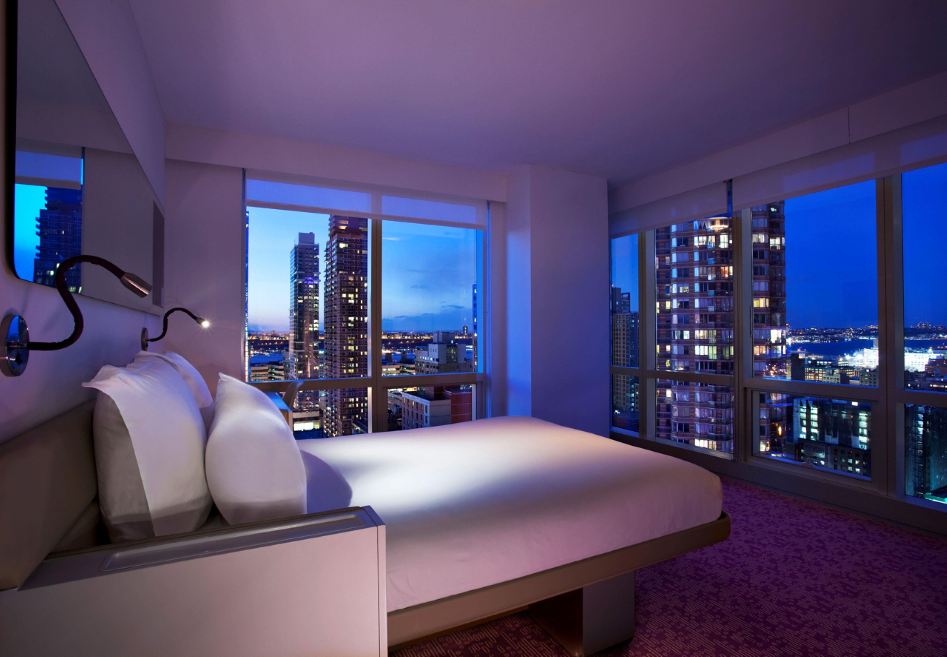 A modern bedroom with a bed, large windows, and a city view.