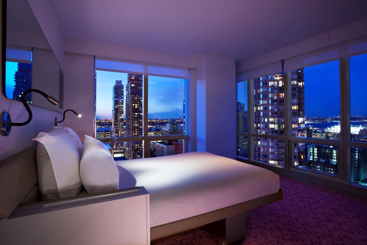 Una camera da letto moderna con un letto, ampie finestre e vista sulla città.