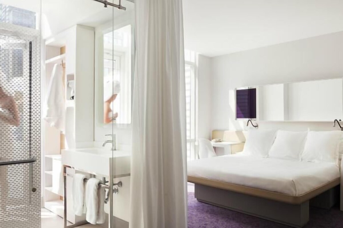 Camera d'albergo con un letto, un comodino, una televisione, uno specchio e un bagno con doccia e lavandino.