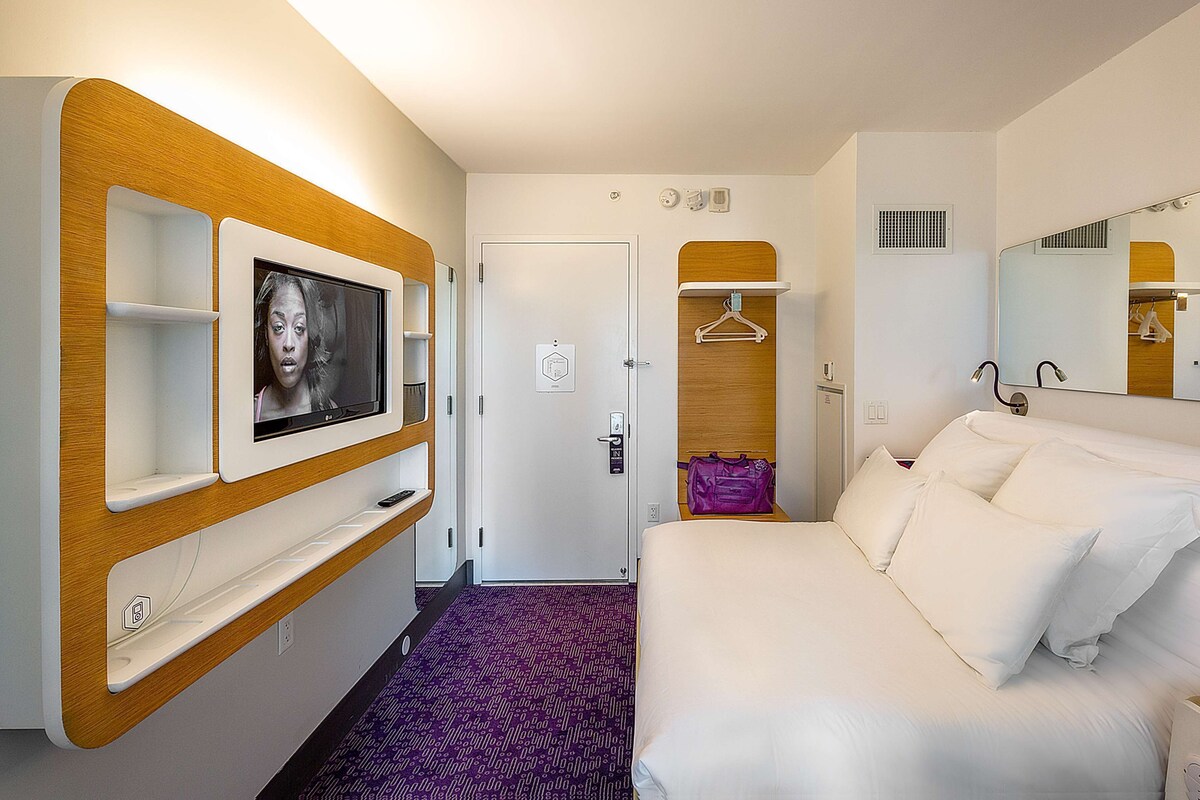 A hotel room with a bed, a TV mounted on the wall, a wardrobe, a mirror, and a door leading to another room.