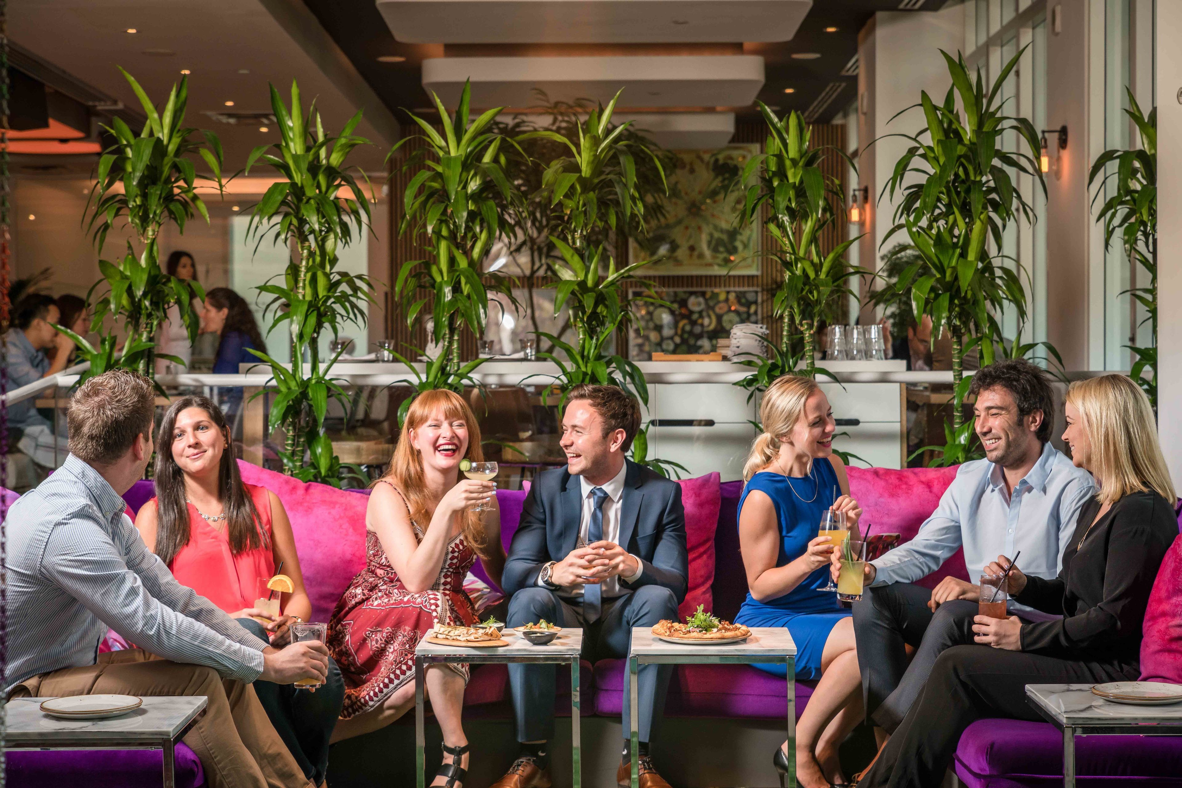 Un groupe de personnes est assis sur un canapé violet dans un restaurant où l’on trouve des tables et des chaises, et l’ensemble est agrémenté de plantes en pot.
