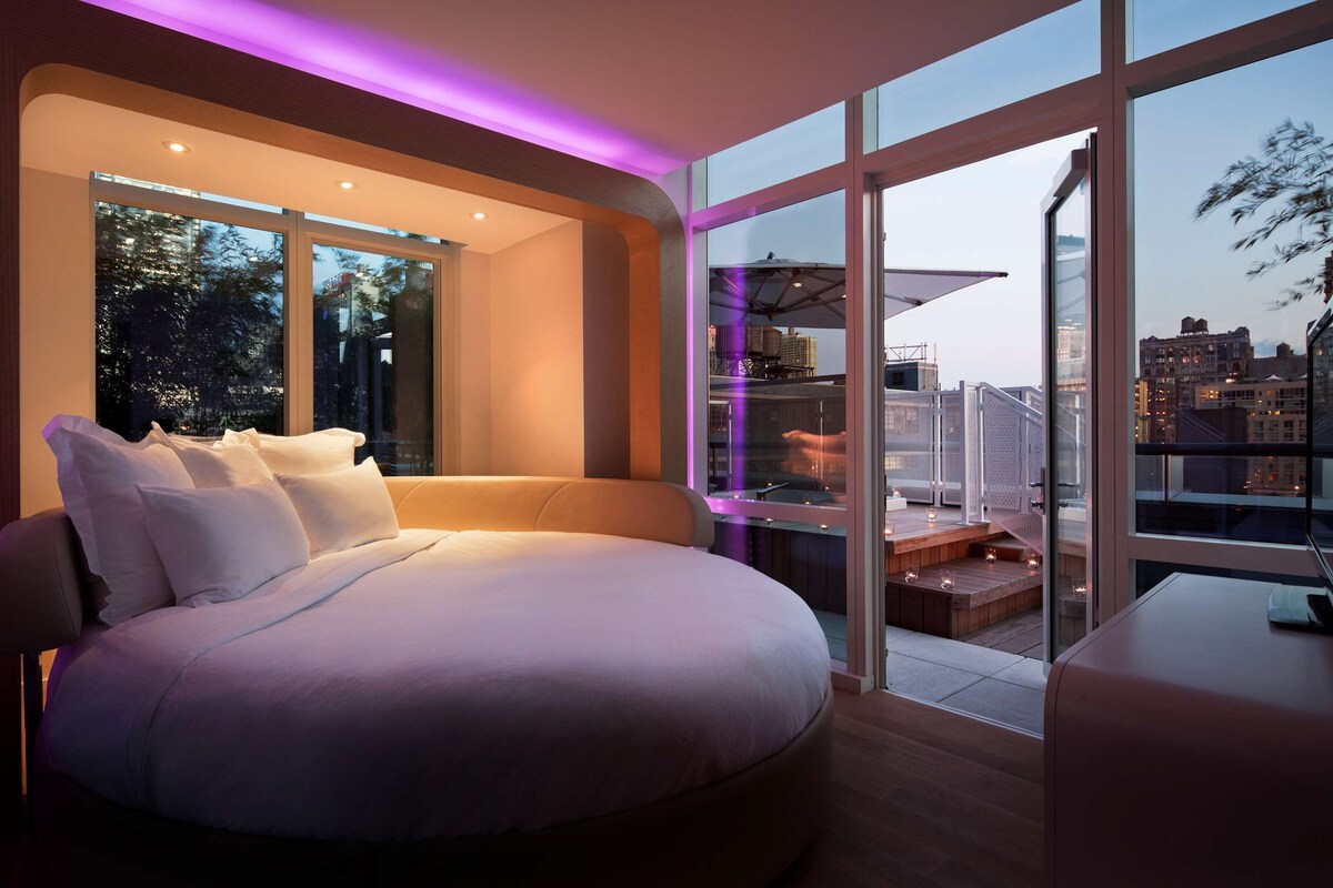 A modern bedroom with a large bed, a view of the city, and a balcony with outdoor seating.