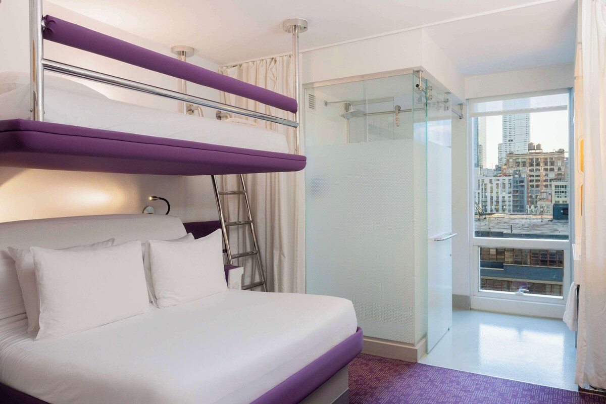 A hotel room with a bunk bed, a glass shower, and a city view.