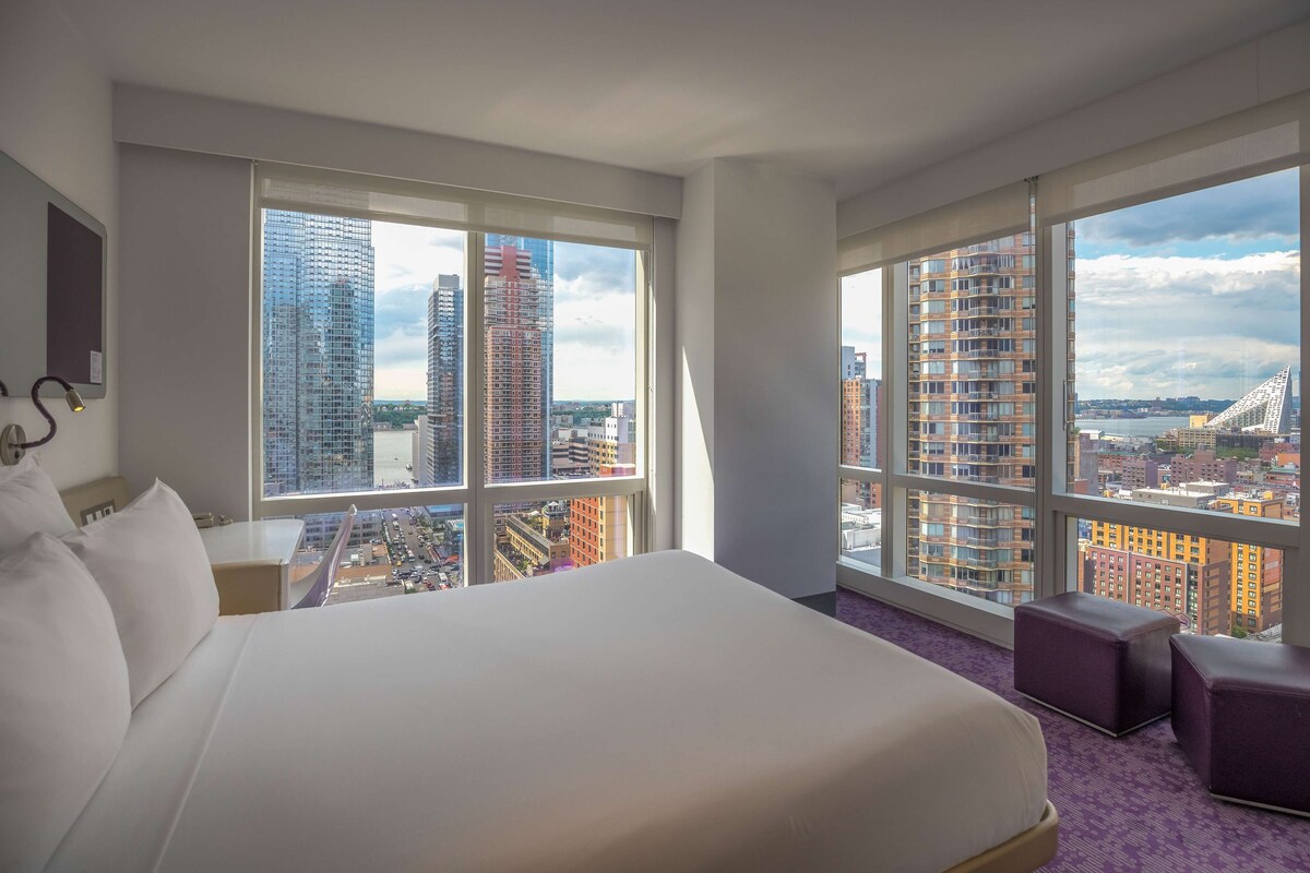 A hotel room with a large bed, two small seating areas, and large windows offering a city view.