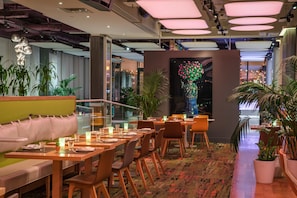 A modern restaurant with wooden tables and chairs, a green cushioned bench, and potted plants.
