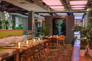 A modern restaurant with wooden tables and chairs, a green cushioned bench, and potted plants.