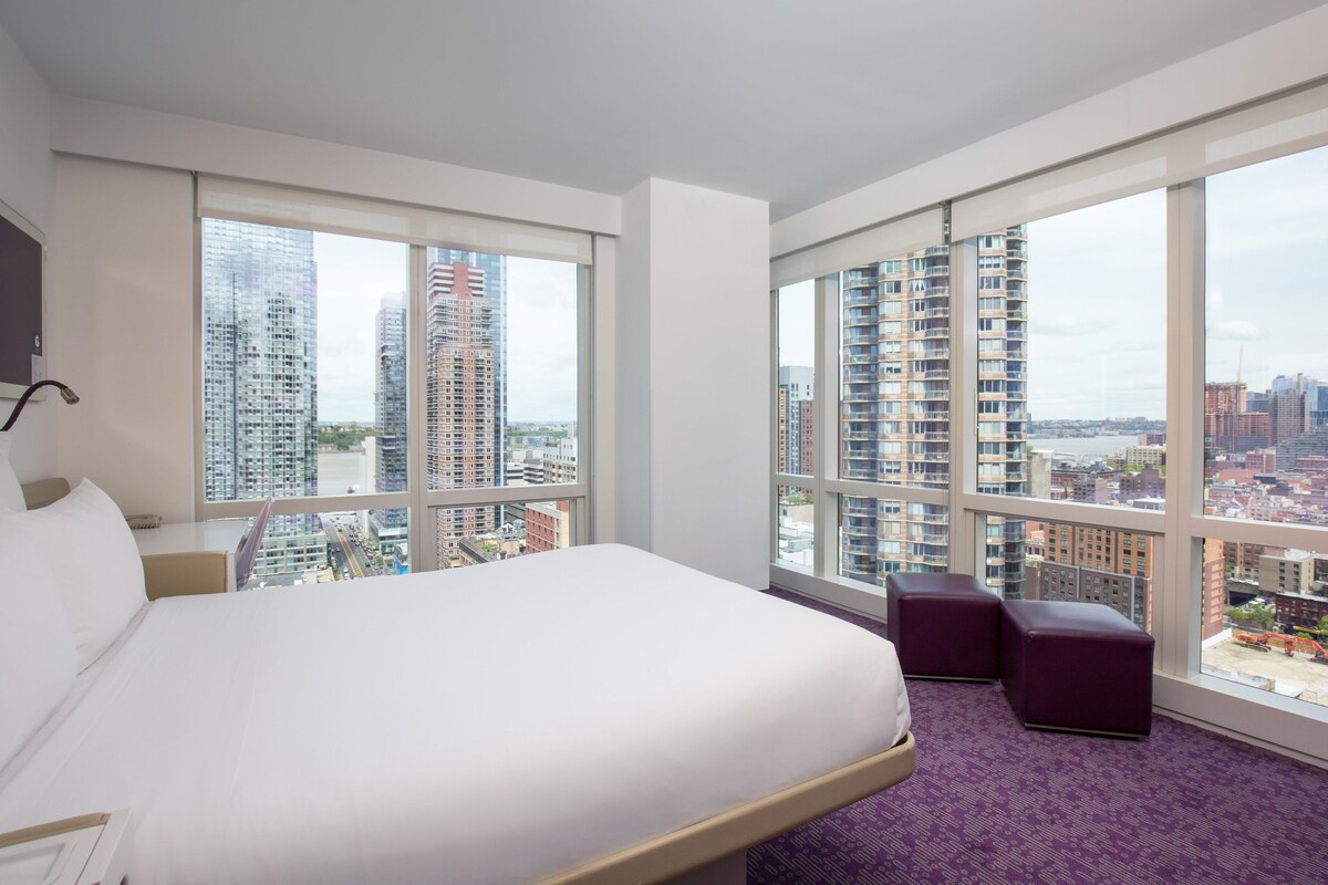 Una camera d'albergo con un letto grande, una scrivania, una sedia e vista sulla città attraverso ampie finestre.