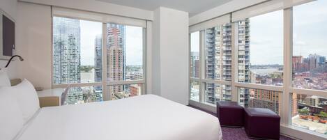 A hotel room with a large bed, a desk, a chair, and a view of a cityscape through large windows.