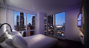 Un dormitorio con una cama grande, una mesita de noche, un televisor y vistas a un paisaje urbano al atardecer.