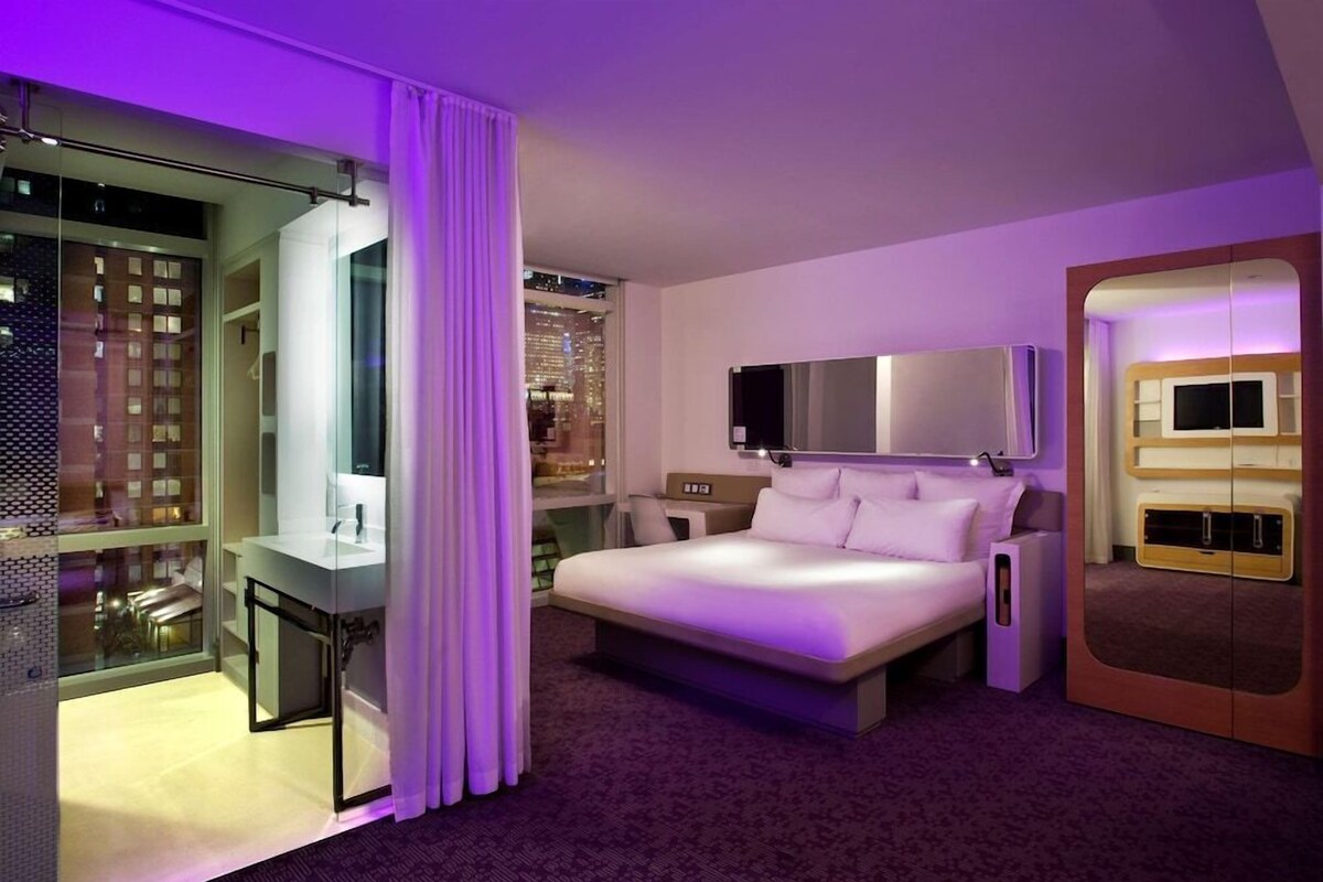 Una moderna camera d'albergo con un letto grande, una televisione, una cabina doccia in vetro e vista sulla città.