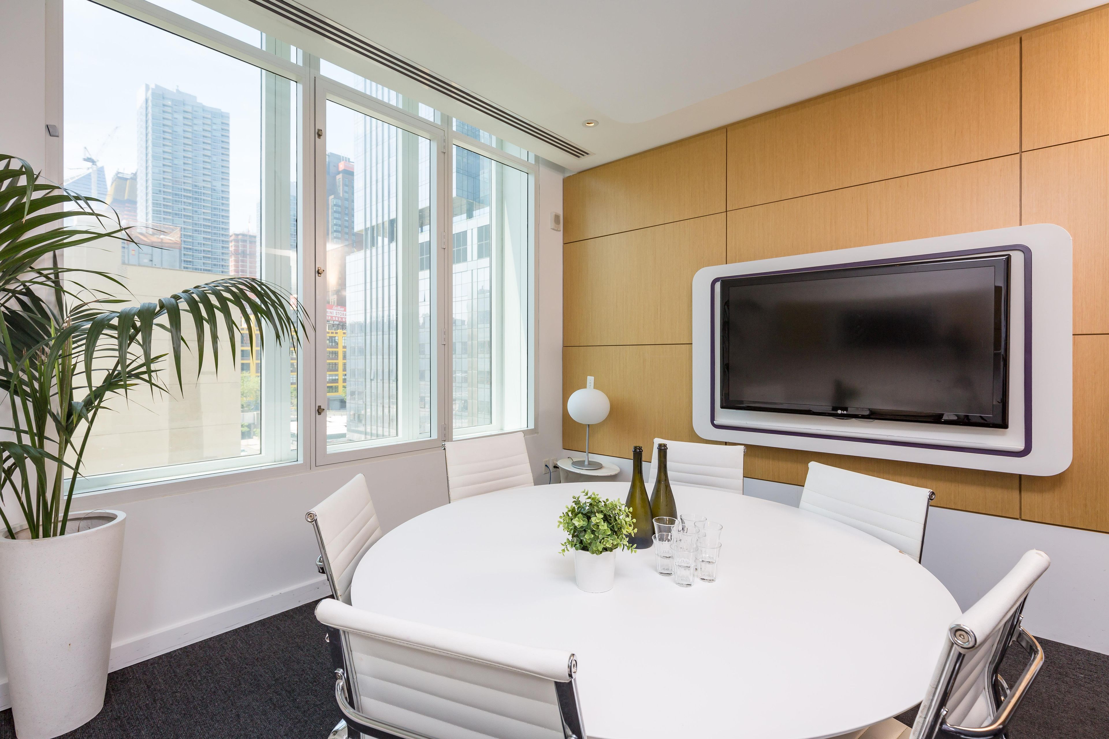 A conference room with a large TV, white chairs, a round table, a potted plant, and a city view through large windows.