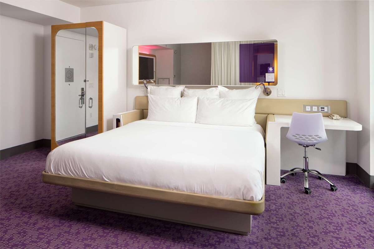 A hotel room with a large bed, a desk with a chair, a mirror, and a wardrobe.