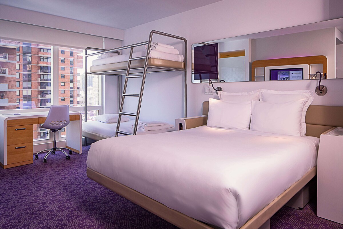 A hotel room with a bunk bed, a desk with a chair, a computer, and a large window with a city view.