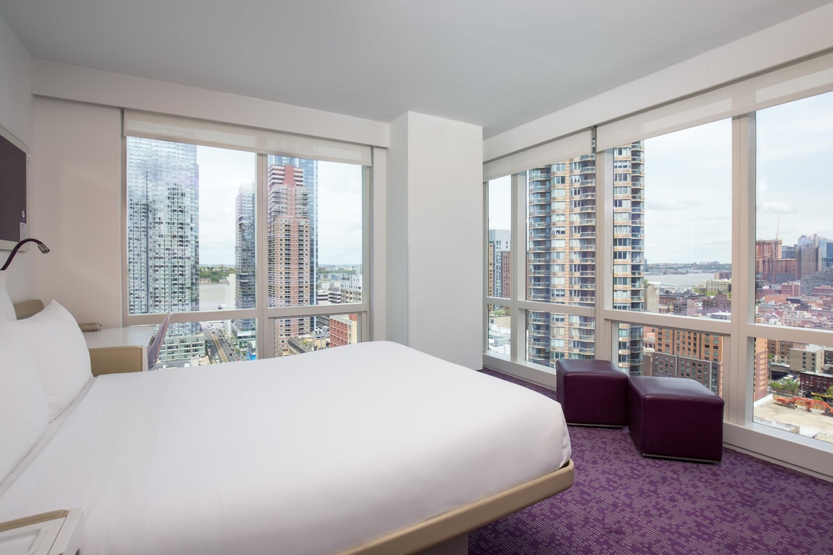 A hotel room with a large bed, a desk, a chair, and a view of a cityscape through large windows.