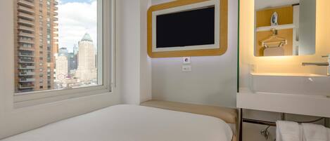 A hotel room with a bed, a window with blinds, a wall-mounted TV, a bathroom with a sink and mirror, and a towel rack.