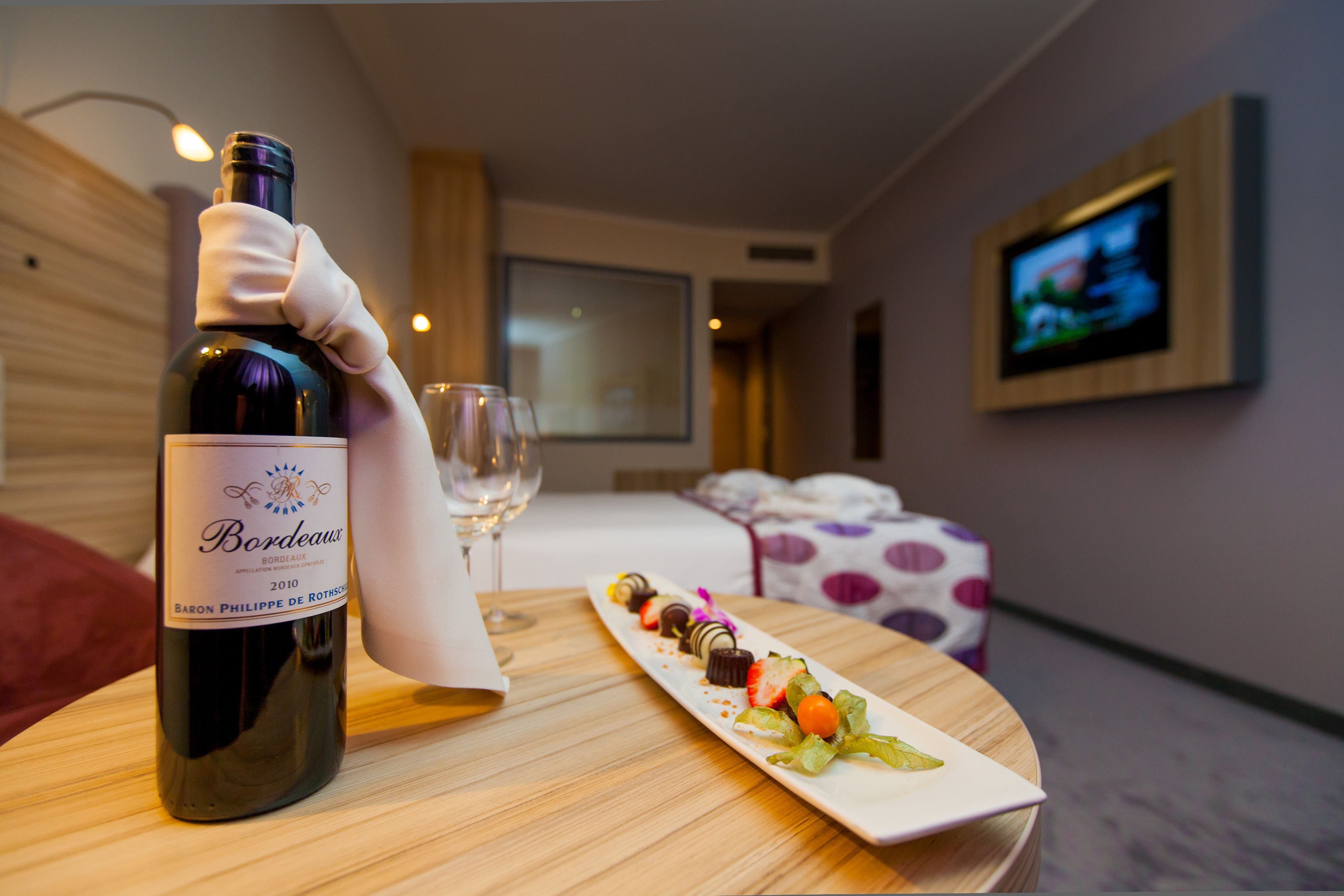 Une bouteille de vin de Bordeaux, un verre et une assiette remplie de chocolats variés, le tout posé sur une table en bois dans une chambre d’hôtel.
