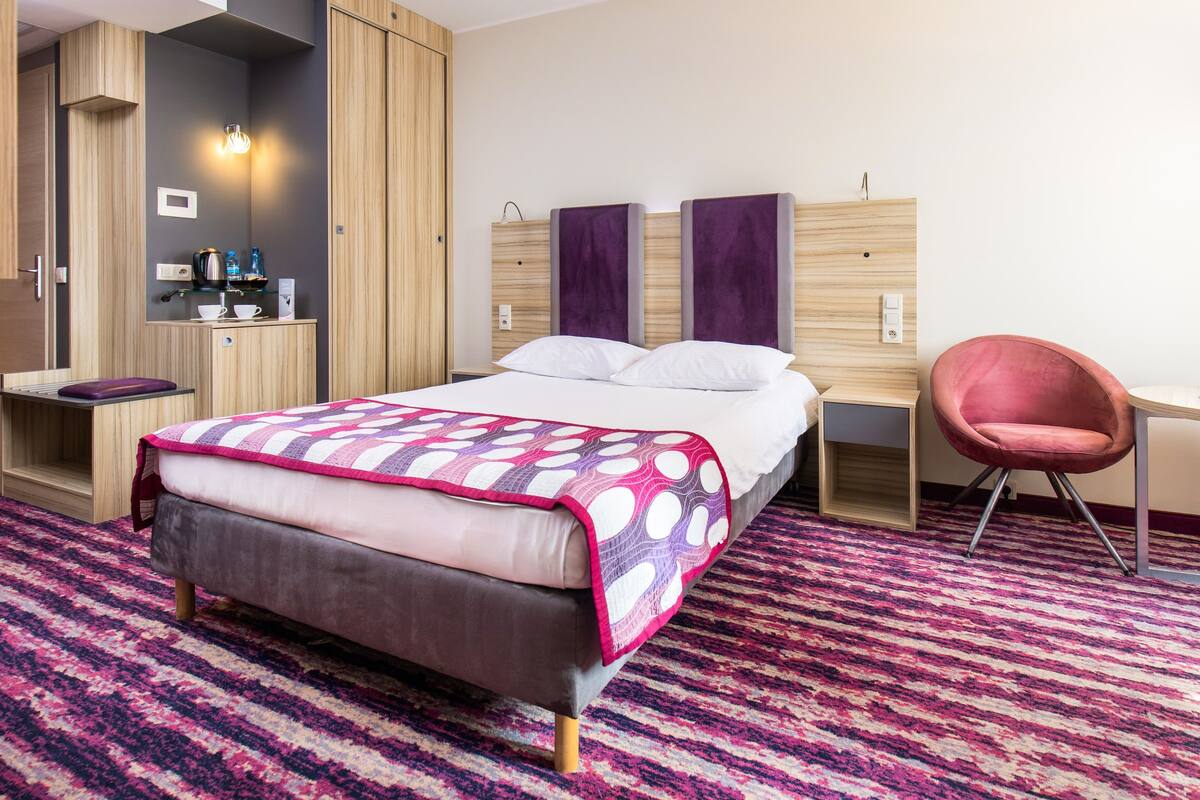 Habitación de hotel con cama, escritorio, silla, armario y un estante pequeño con artículos de aseo.
