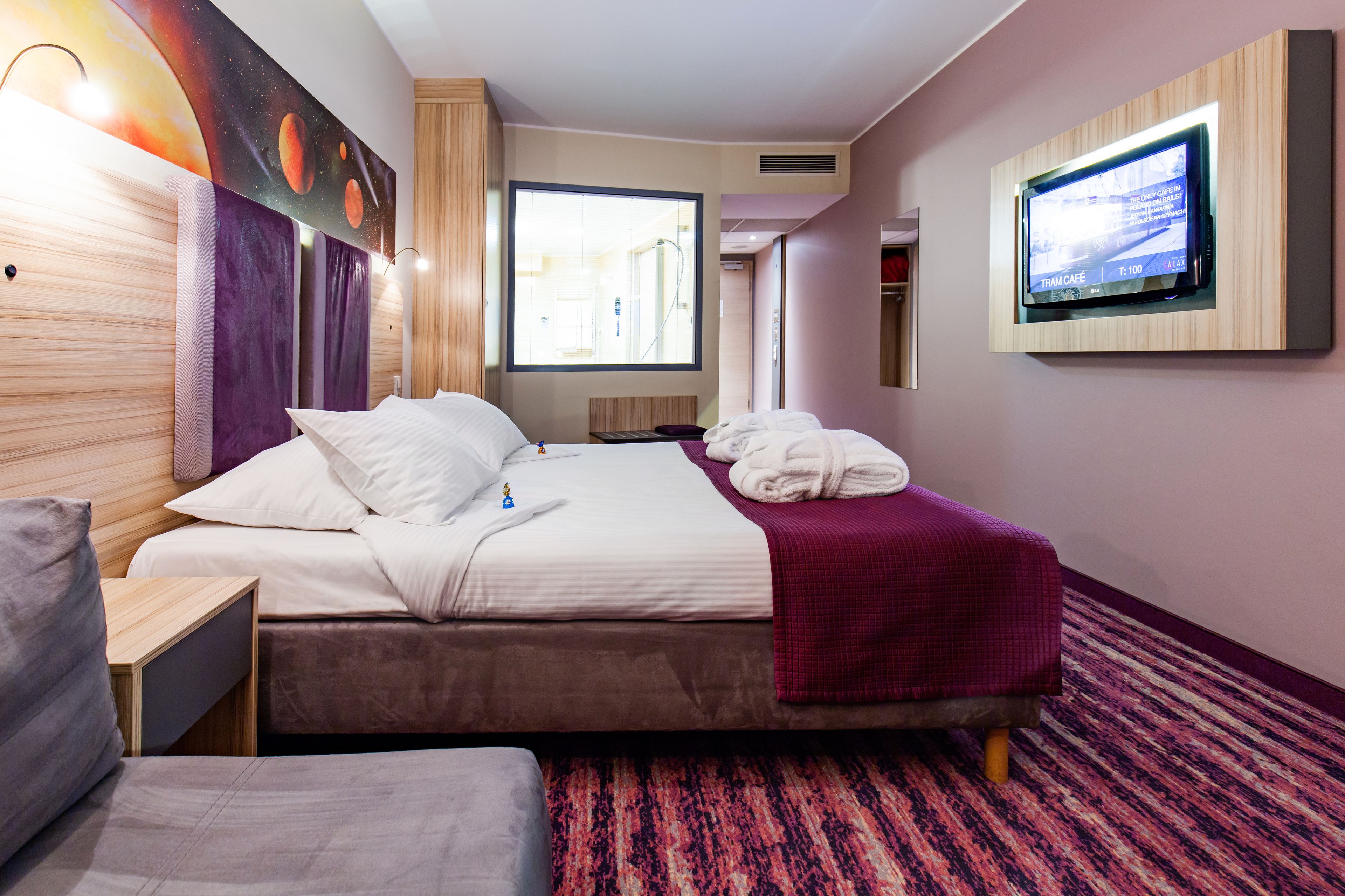 A hotel room with a large bed, a smaller bed, a TV mounted on the wall, and a window providing a view of the hallway.