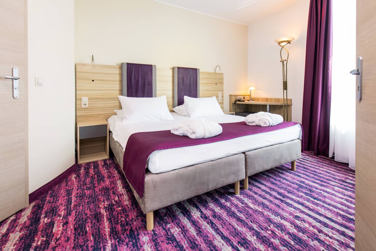 A hotel room with a double bed, a desk, a floor lamp, and a purple curtain.