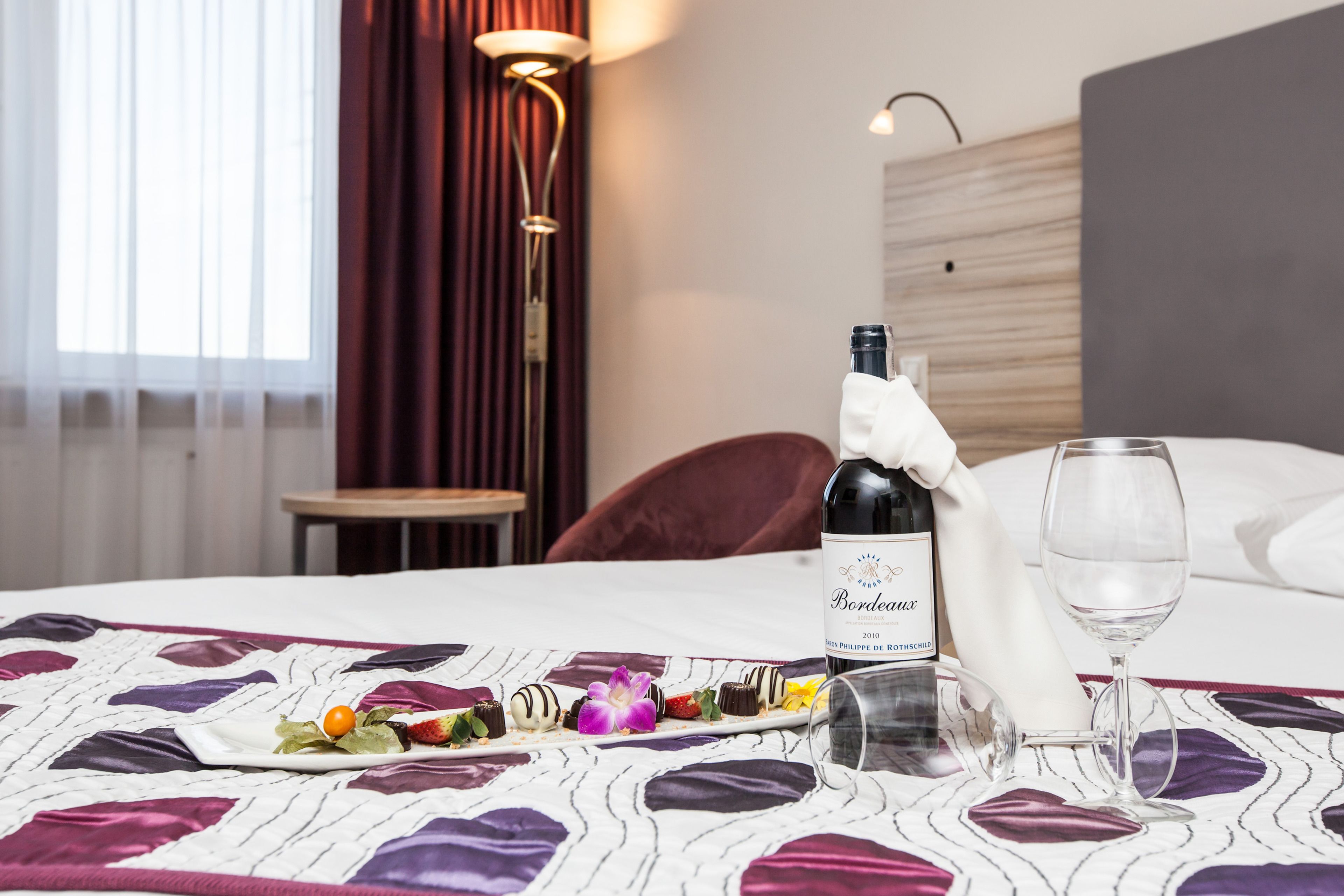 Una habitación de hotel con una cama, una botella de vino de Burdeos, una copa y un plato de bombones.