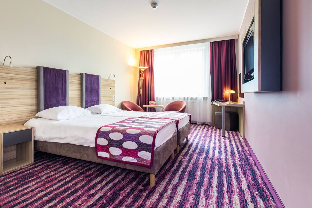 A hotel room with a bed, two chairs, a desk, a lamp, a TV, and a window with curtains.