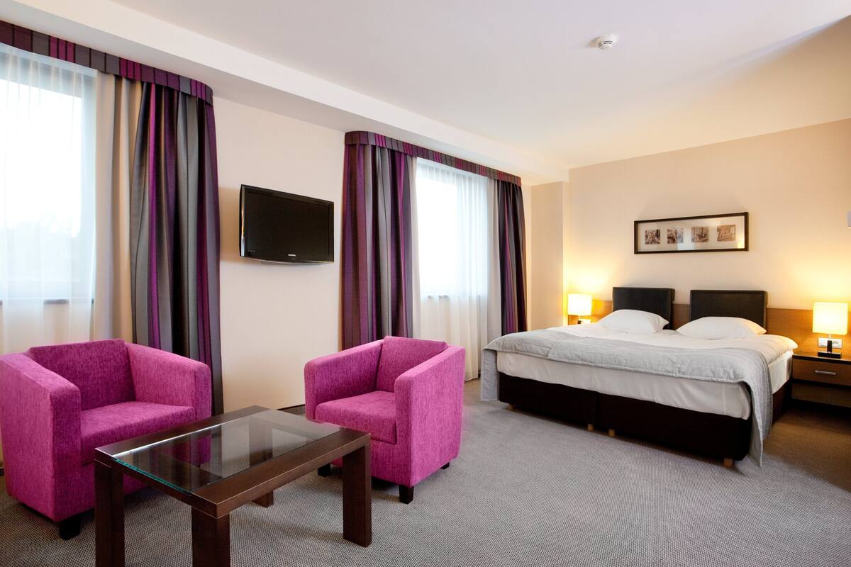 A hotel room with a bed, two chairs, a TV, and a bedside table.